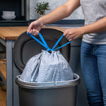Person tying blue drawstrings on a filled gray trash bag inside a kitchen trash can demonstrating Hefty secure-fit closure