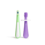 Two toddler training toothbrushes on white background: a purple silicone 360° massaging brush and a green soft-bristled brush.