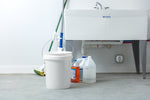 White bucket with lid and handle on a concrete floor next to cleaning supplies under a utility sink