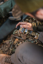 Person seated outdoors holding a Ben's scent free hunting formula spray can with gear and leaves in background