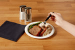 Hand using a black plastic spoon to scoop mashed potatoes from a paper plate with meatloaf and asparagus