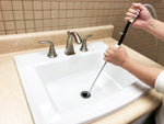 Person holding a manual drain auger with white handles and feeding the coiled cable into a bathroom sink drain