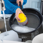 Person using a yellow Scrub Hub to scrub a nonstick frying pan over a sink with soap suds visible