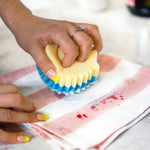 Close-up of a hand using a blue abrasive side of a Scrub Mommy sponge to scrub a red stain on a striped cloth