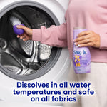 Woman loading a front‑load washing machine while holding a Snuggle Lavender Breeze bottle and a purple scoop of scent booster granules