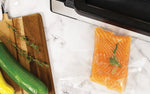 Vacuum-sealed salmon fillet resting next to a vacuum sealer and cutting board with herbs