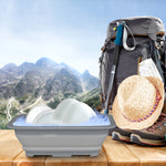 Collapsible gray wash basin with soapy water and dishes in foreground, hiking gear and mountains blurred in background.