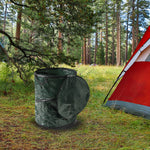 Dark green collapsible trash can shown outdoors beside a tent in a forested campsite scene