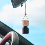 Hanging car diffuser with wooden top and dark bottle suspended from rearview mirror inside a vehicle, soft sky background