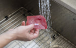 Hand holding a red Lodge silicone scraper under running water at a kitchen sink, water splashing over the tool surface.