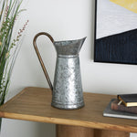Galvanized metal watering pitcher displayed on a wooden table next to artwork and plants