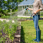 Person watering raised garden beds with spray nozzle attached to a dark textured hose, greenhouse visible in background