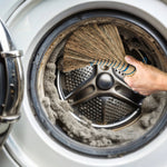Hand holding a straw whisk broom inside a washing machine drum sweeping away lint and debris.