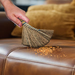 Hand sweeping crumbs from a brown leather sofa with a curved straw whisk broom, crumbs scattered on the seat.