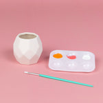Unpainted white geometric planter beside a paintbrush and plastic palette with orange and pink paint wells on pink background