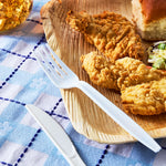 White disposable plastic fork placed beside fried chicken on a wooden plate over a checkered cloth.
