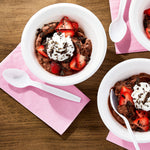 White plastic spoon beside bowls of chocolate dessert and strawberries on a wooden surface with pink napkins