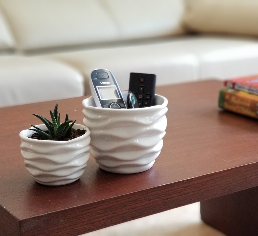 Two white ceramic pots with wavy textured sides on a wooden table, one holding remotes and one a small succulent.