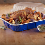 Roasted whole chicken with vegetables served in a blue-lined disposable aluminum roasting pan with lid behind