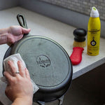 Person wiping the bottom of a Lodge cast iron skillet on a counter with the Lodge seasoning spray and brush visible in the background.