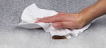 Hand wiping a liquid spill on a textured surface using a white disposable cleaning towel.