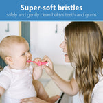 Mother using a pink elephant-shaped training toothbrush to gently brush a baby’s teeth, with headline about super-soft bristles above.