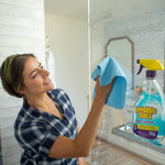 Woman wiping a bathroom mirror with a blue cloth while an Invisible Glass spray bottle sits on a shelf, reflecting in the mirror