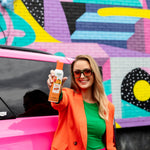 Smiling woman in colorful outfit holding a Citrus Magic Tropical Citrus car spray beside a pink car and mural