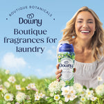 Smiling woman holding a Downy Folia scent booster bottle amid a floral meadow and headline text about boutique fragrances