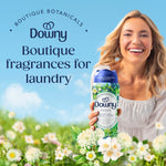Smiling woman holding a Downy Folia scent booster bottle amid a floral meadow graphic and 'Boutique fragrances for laundry' text