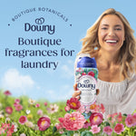 Smiling woman holding a Downy Peonia scent booster bottle against a floral meadow and sky with text about boutique fragrances for laundry