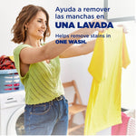 Woman holding up a yellow shirt in a laundry room with bilingual copy claiming the product helps remove stains in one wash