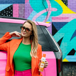 Woman leaning on a pink car holding a can of Citrus Magic Tropical Citrus car spray in front of a colorful mural
