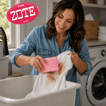 Smiling woman scrubbing a muddy shirt at a sink with a pink Zote soap bar, laundry room and washing machine visible in background.
