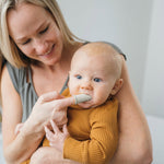 Woman holding a baby and using a gray silicone finger toothbrush to gently clean the infant's mouth