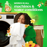 woman hugging white towels by a front-load washer with a gain fabric softener bottle on top and tropical flowers on a green background