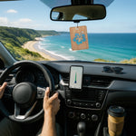 Wooden rectangular hanging air freshener with blue hibiscus cutout suspended from a car rearview mirror over a coastal drive.