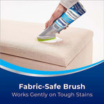 Hand using Woolite foam cleaner with green brush on a light fabric ottoman, graphic text states fabric-safe brush
