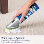 Hand-held Woolite spray with built-in brush applying foamy cleaner to a light carpet near a wooden furniture leg.