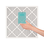 Hand placing a turquoise Yankee Candle hanging air freshener onto a square furnace or A/C filter against a white background.