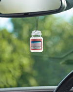 Yankee Candle Pink Sands jar air freshener suspended from a rearview mirror with blurred green foliage background