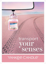 Yankee Candle Pink Sands car jar air freshener hanging from a rearview mirror over a pink-sand beach background