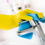 Gloved hand wearing a yellow rubber glove wiping a chrome faucet with a blue cleaning cloth