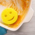 Yellow smile-shaped scrub sponge resting inside a soiled ceramic baking dish showing food residue on a countertop