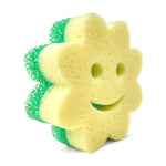 Angled view of the yellow sponge side of a flower-shaped Scrub Mommy with green scrubber layer and smiley face cutouts