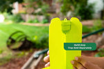 Person holding the bright yellow trap showing a green lure cartridge seated at the trap's top in an outdoor garden scene