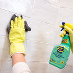 Gloved hand wiping tiled wall with cloth while holding a bottle of Zep Home Pro foaming bathroom cleaner