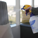 Person spraying a Zep-branded squeeze spray bottle indoors, mist visible from the nozzle toward a seating area