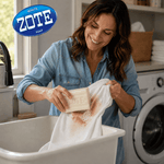 Smiling person at a laundry sink scrubbing a stained white garment with a Zote soap bar, with washing machine and Zote logo visible.